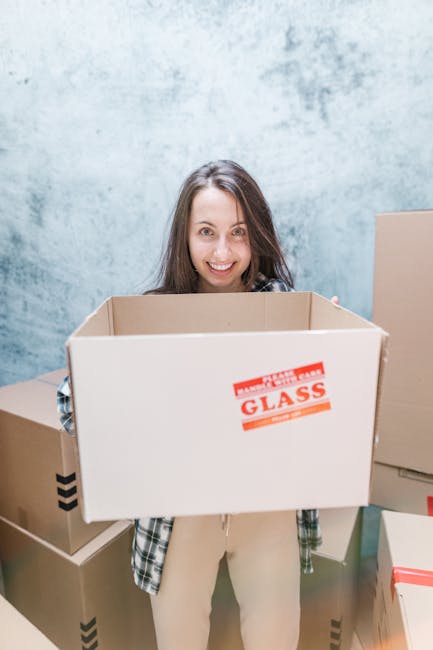 A professional mover wearing a navy blue jumpsuit and a black bandana is inside a property, standing next to a large cardboard box and writing on a clipboard. There are several packing materials and b